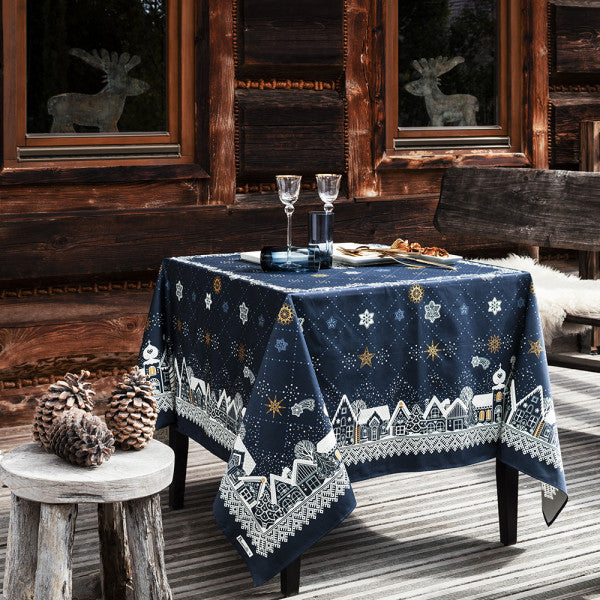 Elegant Beauville Nuit Etoilee Blue tablecloth drapes over a rustic wooden table, featuring winter village and star motifs.