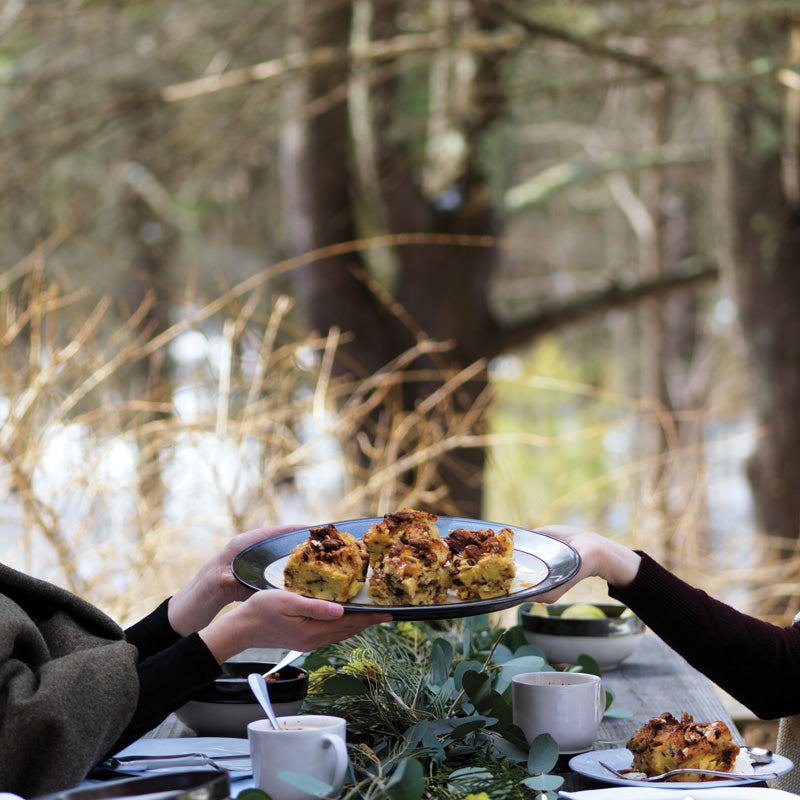 A person serves a footed soup bowl filled with food at an outdoor table surrounded by trees, creating a cozy atmosphere.