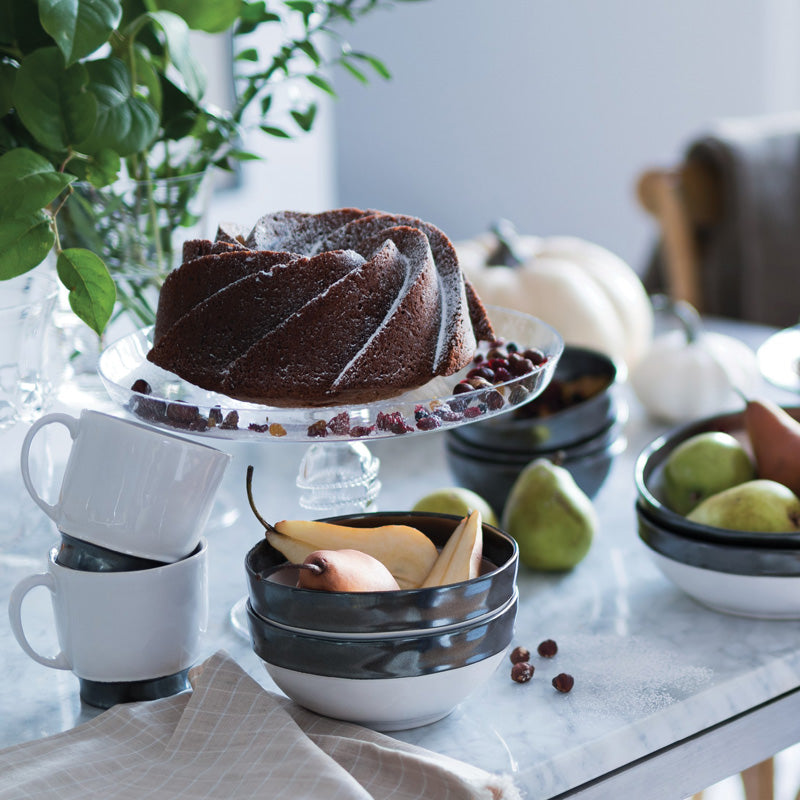 Juliska Pewter Stoneware footed soup bowl displayed on a table with fruit and a cake, surrounded by greenery and decorative items.